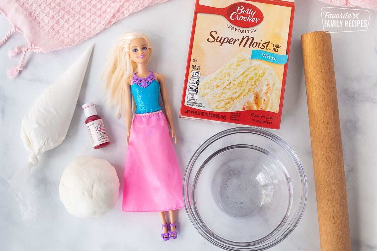 Ingredients to make a Barbie Cake including cake mix, barbie, glass bowls, rolling pin, fondant, frosting and gel coloring.