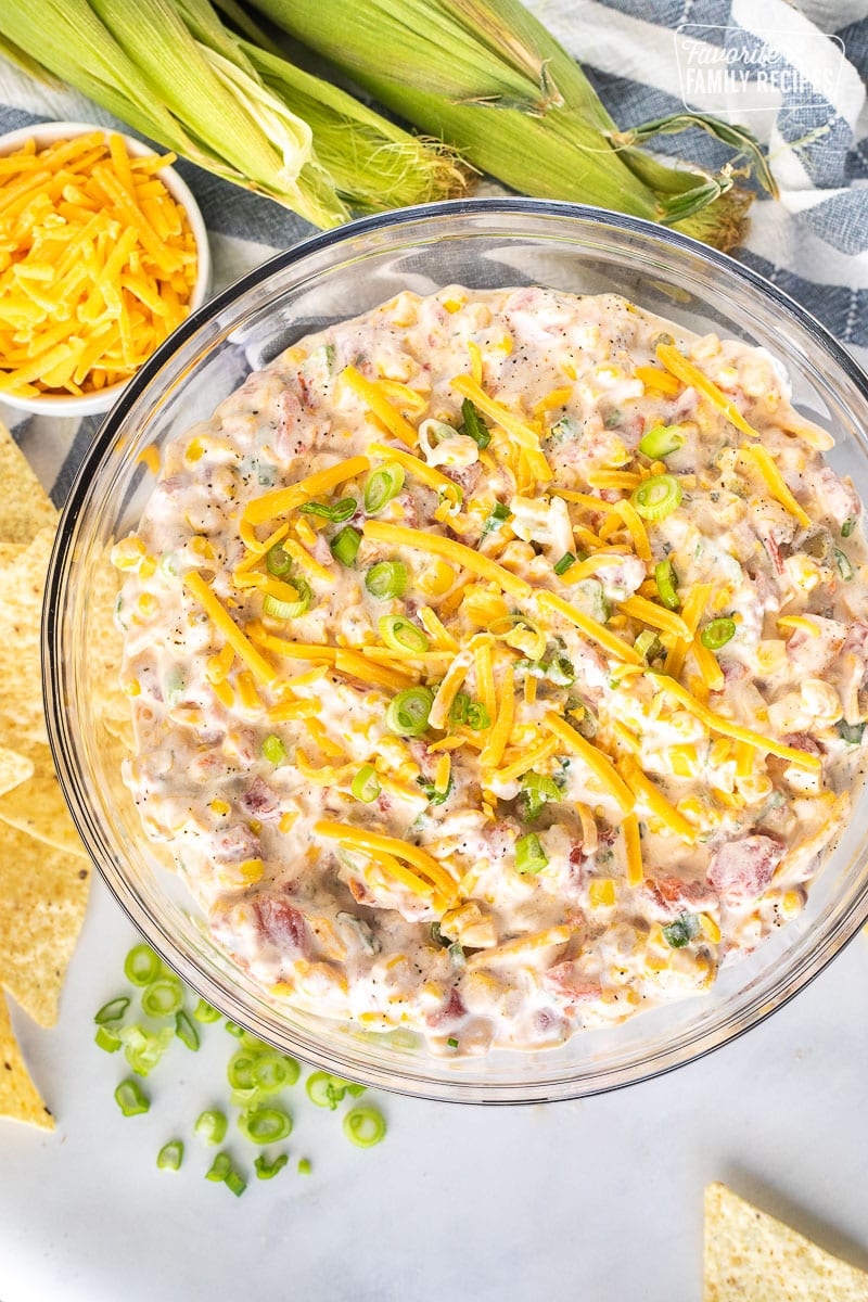 Bowl of Cream Corn Dip next to cheddar cheese, tortilla chips, green onion and corn.