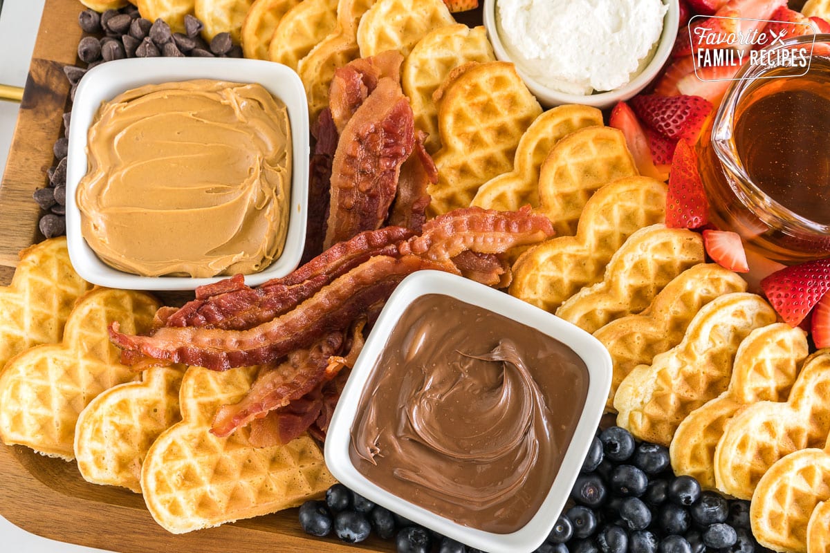 Close up of a breakfast charcuterie board with waffles, bacon, peanut butter, nutella, blueberries and chocolate chips
