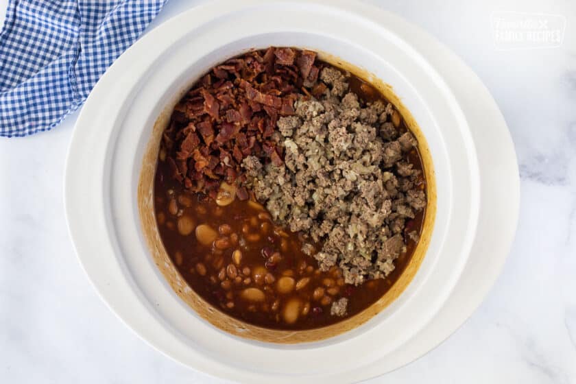 Crockpot with beans mixture topped with cooked bacon and ground beef for Baked Beans.