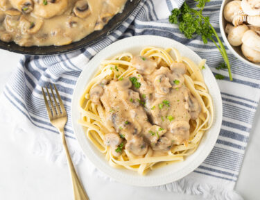 Chicken Marsala over fettuccine pasta on a plate next to a fork and garnished with parsley.