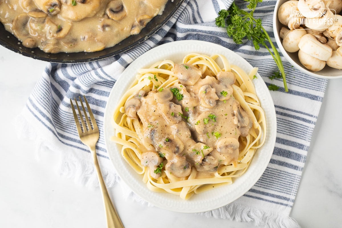 Chicken Marsala over fettuccine pasta on a plate next to a fork and garnished with parsley.