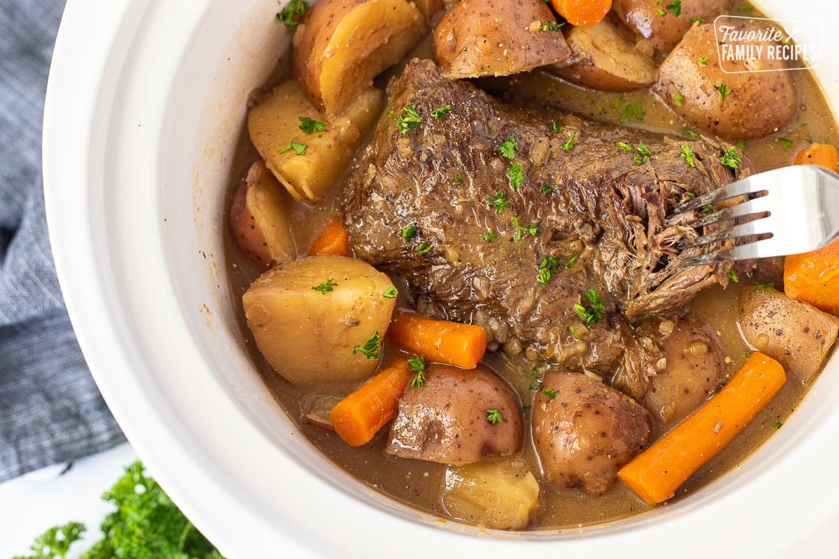 Fork shredding Crockpot Roast Beef surrounded by cooked carrots and red potatoes.