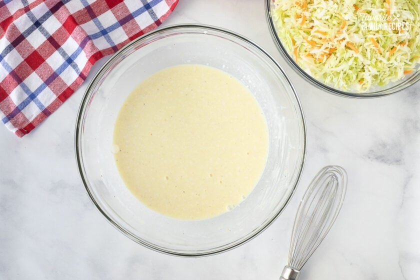 Glass bowl of dressing on Homemade Coleslaw. Whisk on the side.