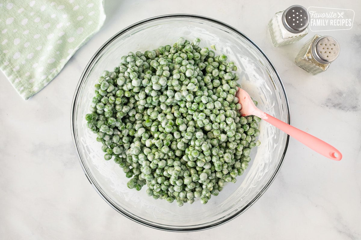 Glass bowl of combined dressing and green peas with spatula with salt and pepper.