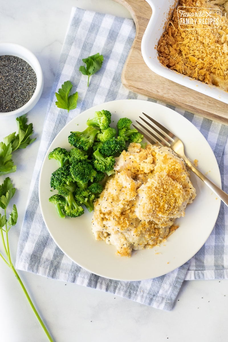 Poppy Seed Chicken Casserole is a tangy, filling meal that the whole family will love! Tender chicken, crispy crust and a sauce to die for. Perfect for busy weeknights. Serving of Poppy Seed Chicken on a plate next to broccoli. Parsley on the side.