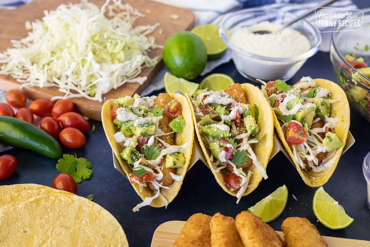 Three Easy Fish Tacos on a taco stand topped with avocado, tomatoes, cilantro, cream sauce and cotija cheese.