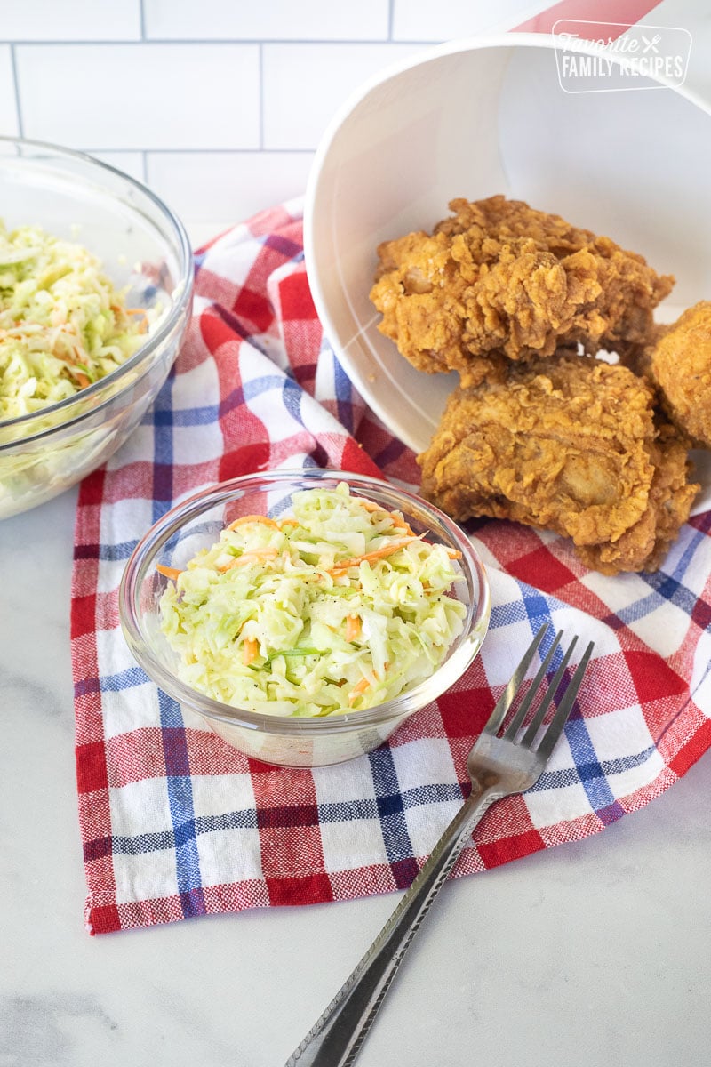 Coleslaw in a small bowl with a fork. Fried chicken and a bowl of extra coleslaw on the side.