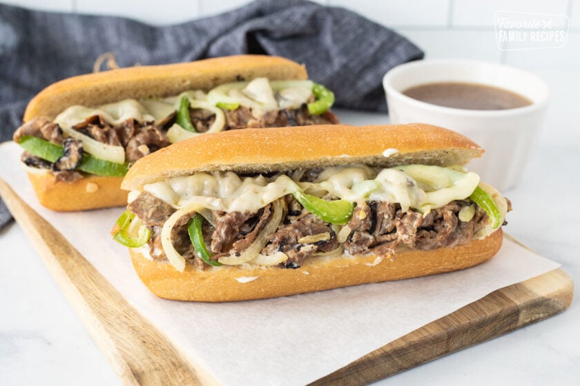 Two Philly Cheesesteak with melted provolone cheese on a cutting board with au jus in a bowl on the side.