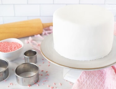 Cake covered with a sheet of fondant on a cake stand. Rolling pin, sprinkles and cutters on the side.