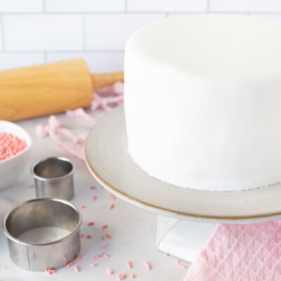 Cake covered with a sheet of fondant on a cake stand. Rolling pin, sprinkles and cutters on the side.