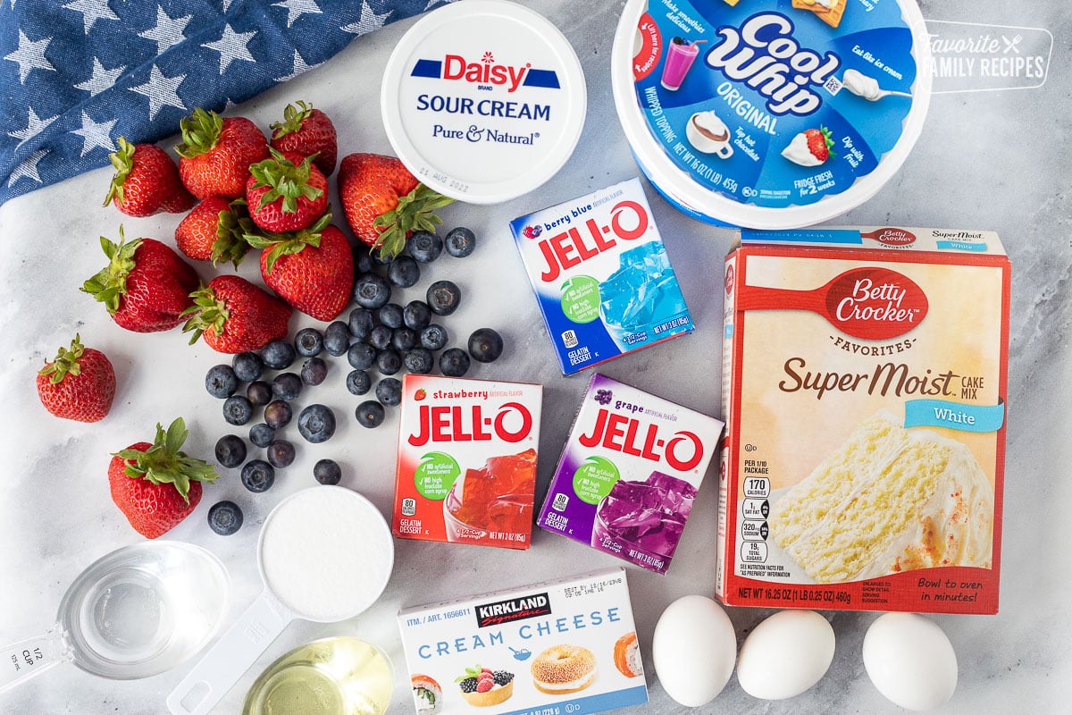 Ingredients to make 4th of July Cake including white cake mix, cool whip, sour cream, blueberries, strawberries, eggs, sugar, oil, water, strawberry jello, berry blue jello and grape jello.