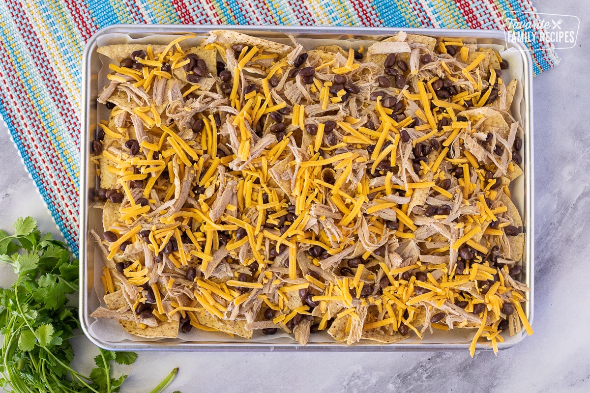Baking sheet with tortilla chips, black beans, shredded pork and shredded cheddar cheese for Pulled Pork Nachos.