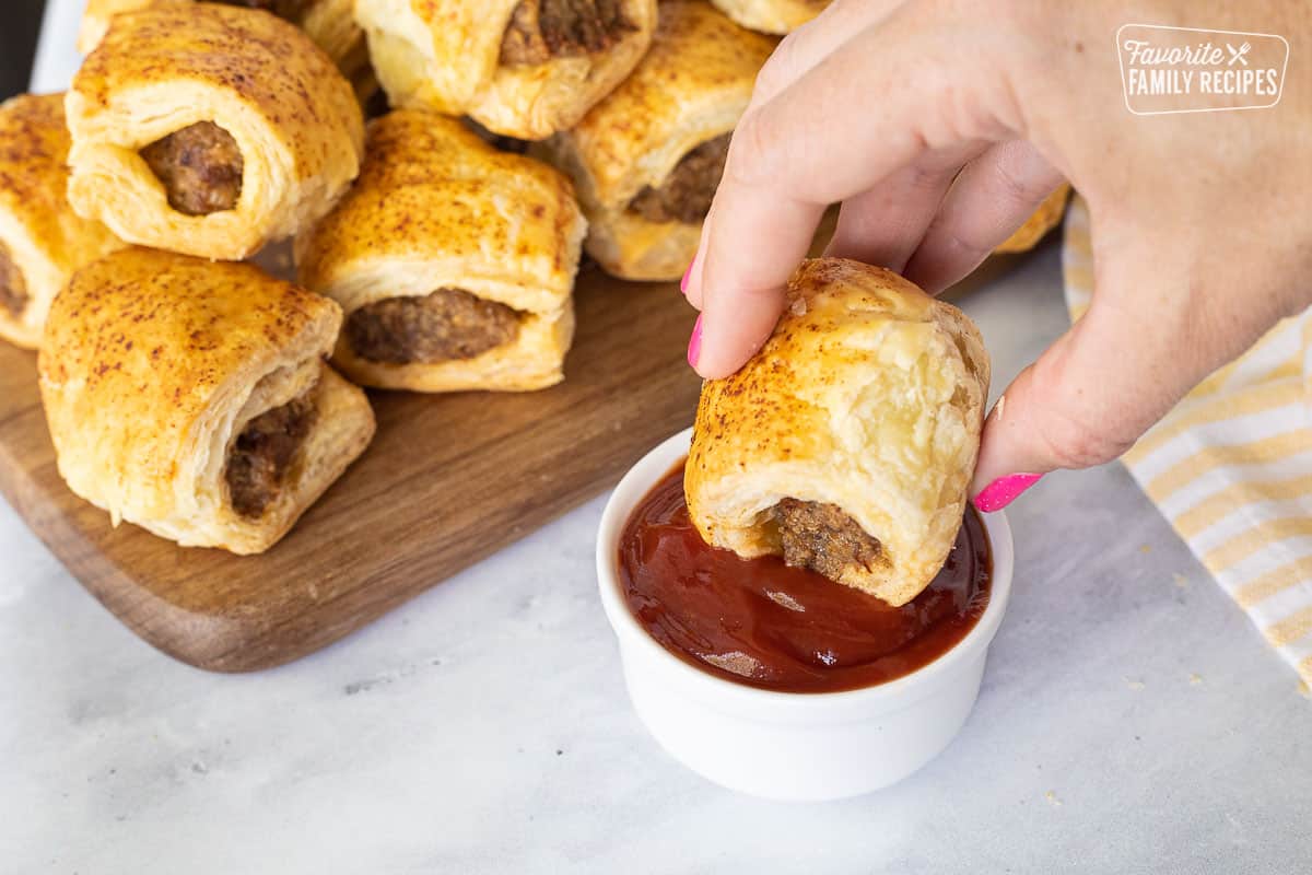 Sausage Rolls are a seasoned, fresh sausage wrapped in a flaky, buttery pastry. They are delicious for breakfast, lunch, or dinner, or as an appetizer. Hand dipping a Sausage Roll in a small bowl of ketchup.