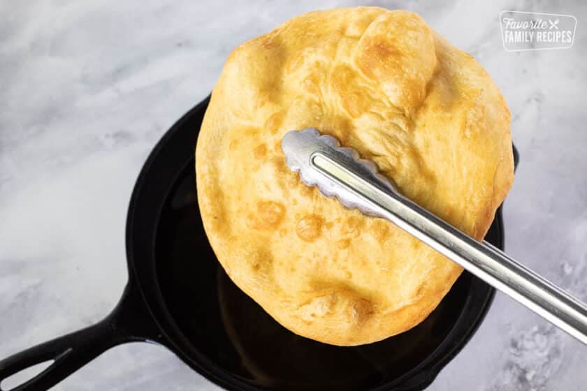 Tongs lifting a piece of Fry Bread from a skillet with oil.