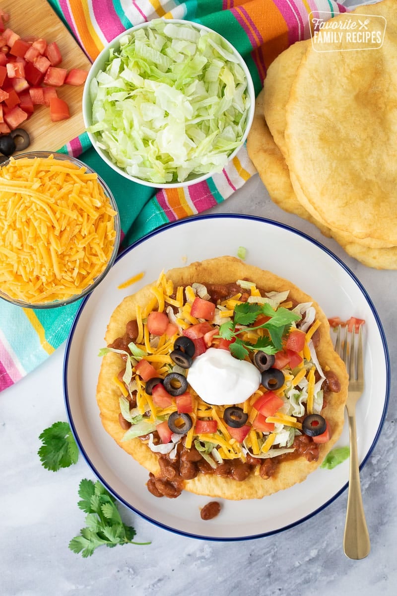 Navajo Taco on a plate next to fry bread, lettuce, cheese, tomatoes and olives.