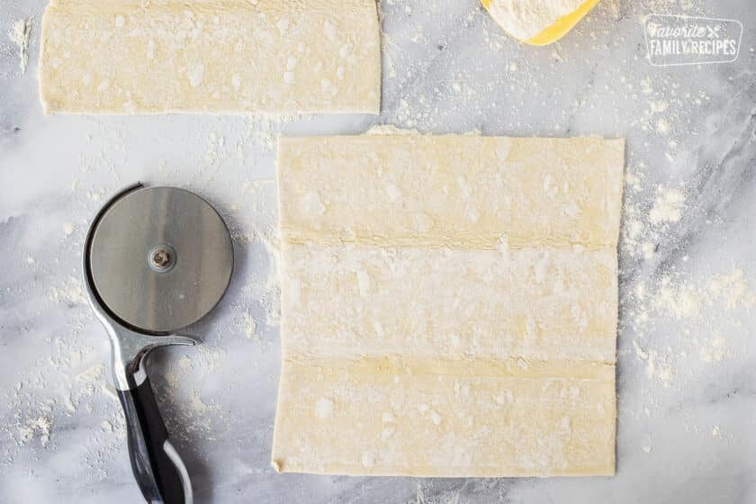 Square puff pastry on a floured surface with pizza cutter for Sausage Rolls.