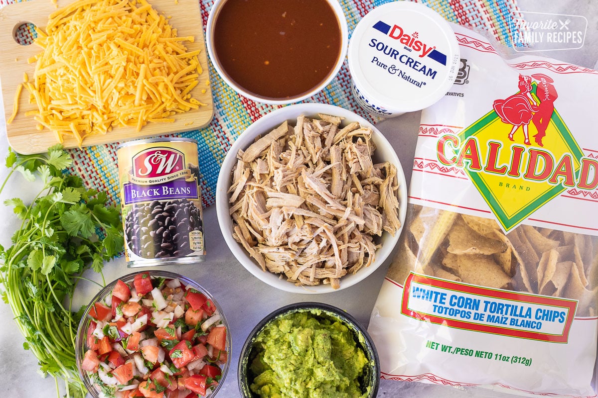 Ingredients to make Pulled Pork Nachos including BBQ sauce, cheddar cheese, sour cream, black beans, cilantro, pico de Gallo, guacamole, shredded pork and chips.