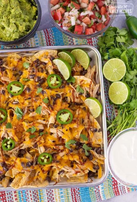 Baking sheet with Pulled Pork Nachos and sliced limes. Pico, guacamole and sour cream on the side.