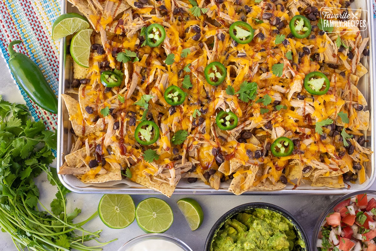 Pulled Pork Nachos on a baking sheet with fresh limes, cilantro, sour cream, guacamole and pico on the side.