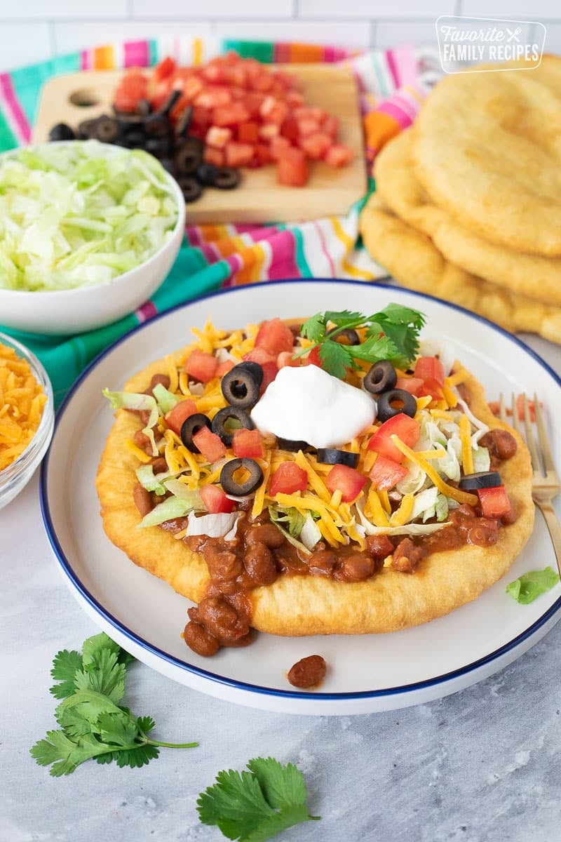 Navajo Taco on a plate made with chili, lettuce, tomatoes, olives, cheese and sour cream.