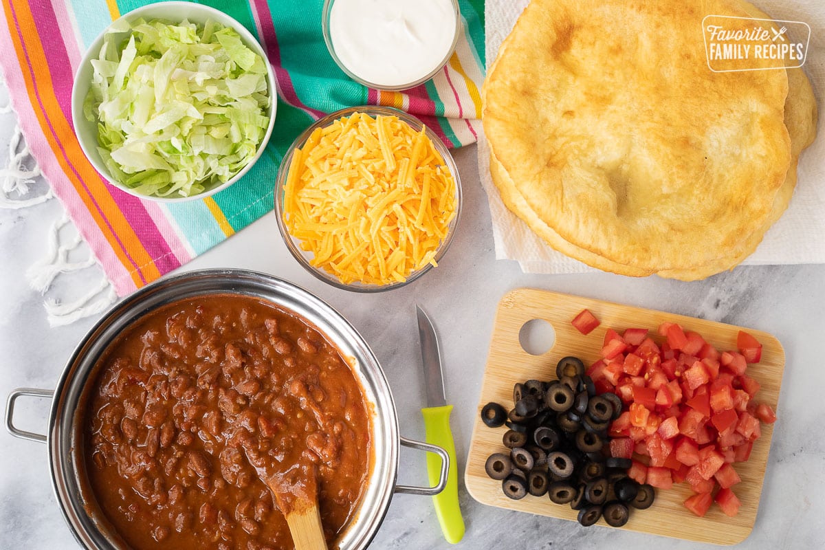 Fry bread next to toppings of cheddar cheese, sour cream, lettuce, tomatoes, olives and chili to make Navajo Tacos.