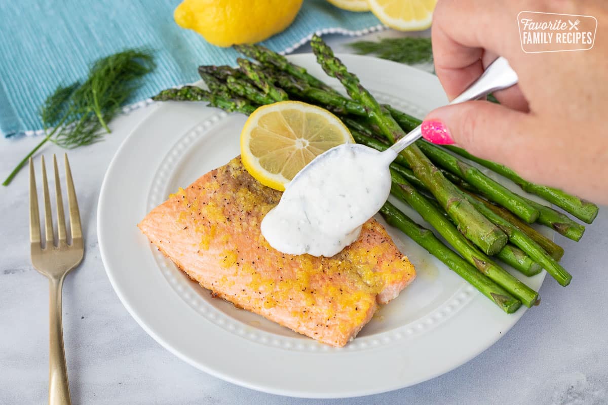 Spooning Tzatziki sauce on top of a fillet of Baked Lemon Salmon on a plate with asparagus.