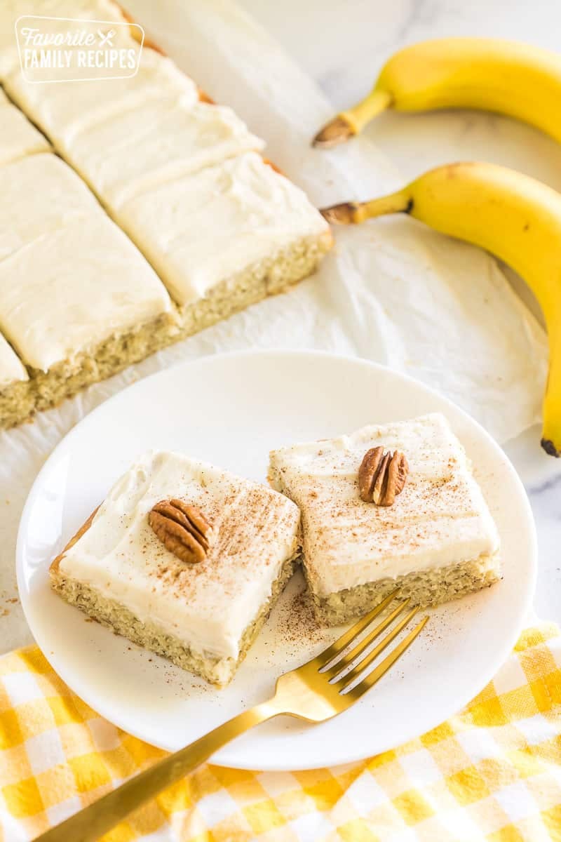 This Banana Cake is the best way to use up those bananas on your counter. The cake comes out so moist and the cream cheese frosting is amazing! Two slices of banana cake on a plate topped with pecans and ground cinnamon.