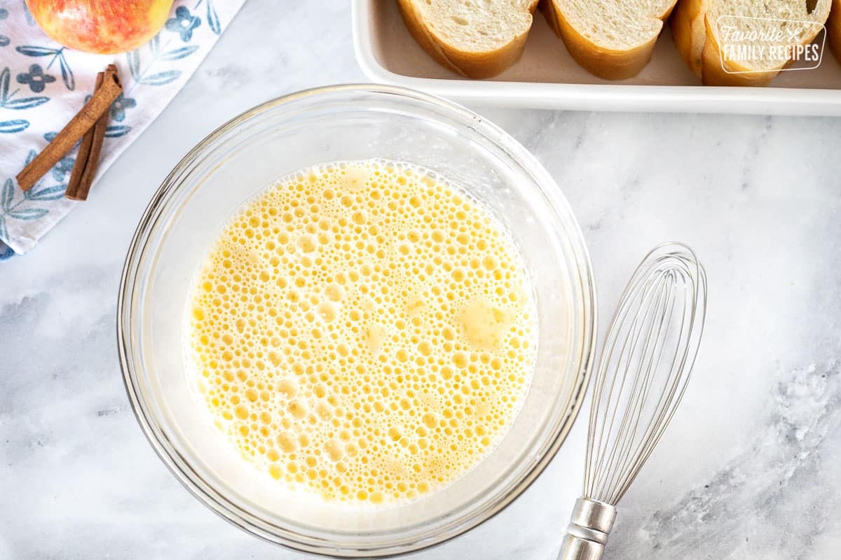 Egg mixture in a mixing bowl next to a whisk for Caramel Apple French Toast Casserole.