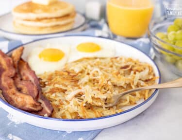 Hash Browns on a fork resting on a breakfast plate with eggs and bacon.