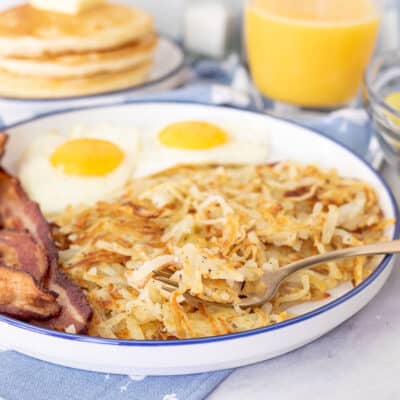 Hash Browns on a fork resting on a breakfast plate with eggs and bacon.