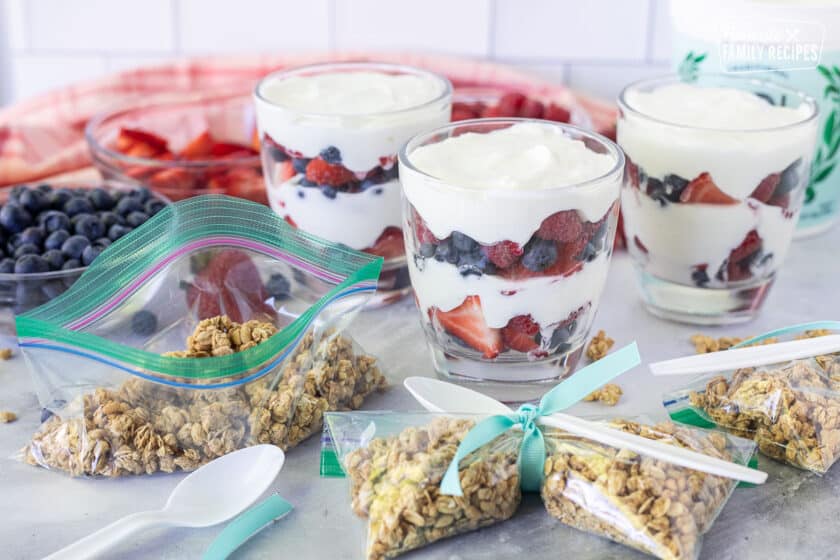 Three make ahead Breakfast Parfaits in cups next to bags of granola and a plastic spoon.