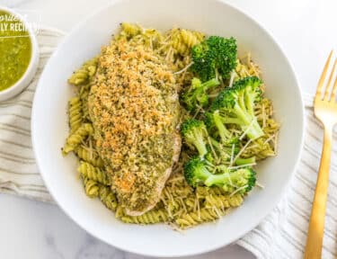 A bowl of pasta with a piece of pesto chicken on top and broccoli