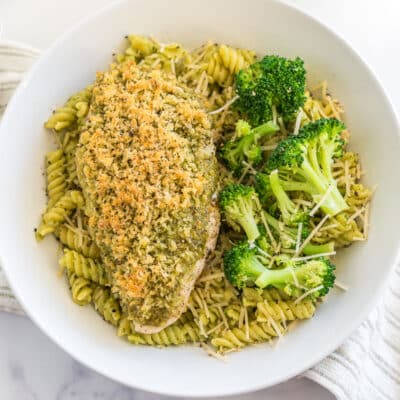 A bowl of pasta with a piece of pesto chicken on top and broccoli