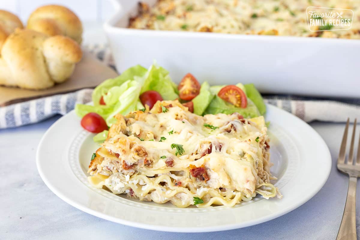 Plate with White Chicken Lasagna piece and a side salad. Bread knots and baking dish of White Chicken Lasagna on the side.