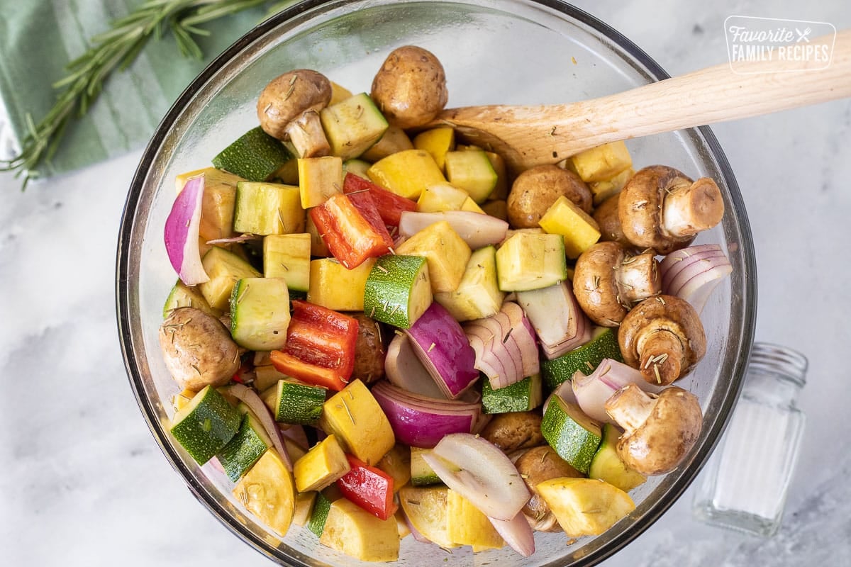 Mixing rosemary and salt in a mixing bowl with fresh cut up vegetables for Oven Roasted Vegetables.