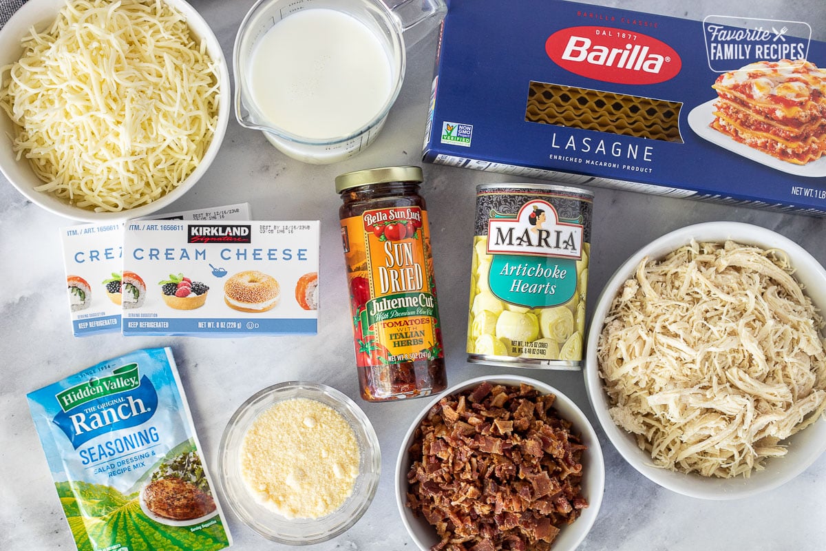 Ingredients to make White Chicken Lasagna including lasagne noodles, mozzarella cheese, milk, cream cheese, sun dried tomatoes, artichoke hearts, shredded chicken, bacon, parmesan cheese and ranch seasoning.