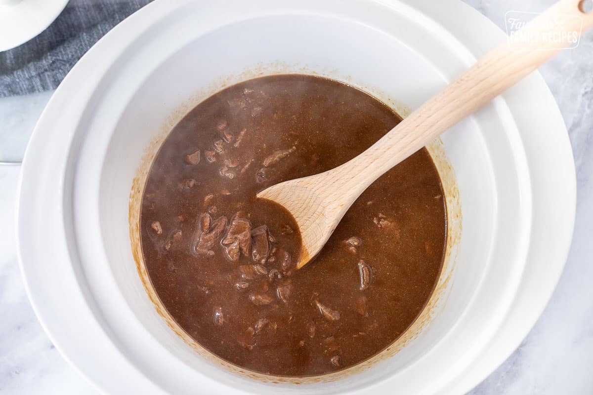 Crockpot with steamy hot steak and gravy and wooden spoon.