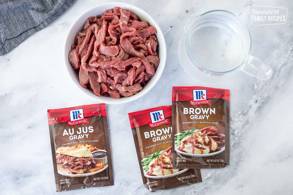 Ingredients to make Crockpot Steak and Gravy including brown gravy mix packets, au jus gravy mix packet, water and thinly sliced top sirloin steak.