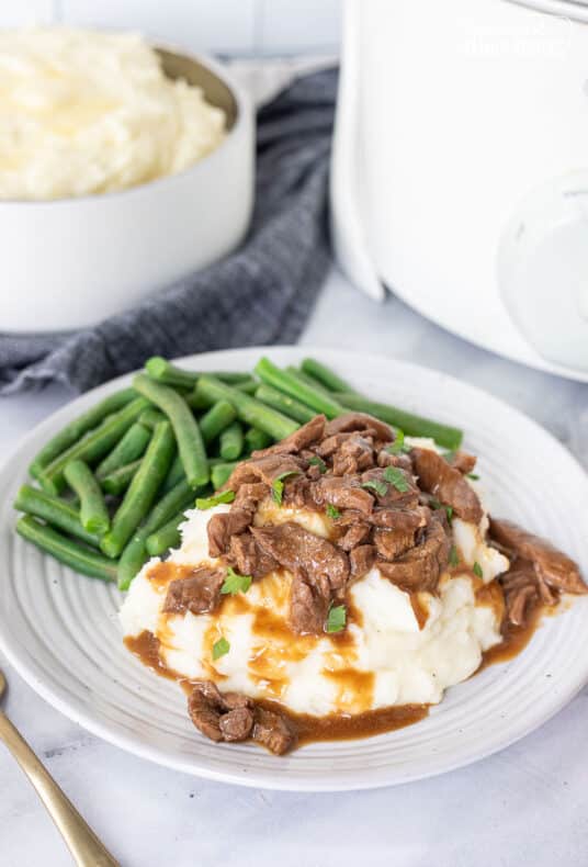 Plate with Crockpot Steak and Gravy over mashed potatoes and green beans on the side.