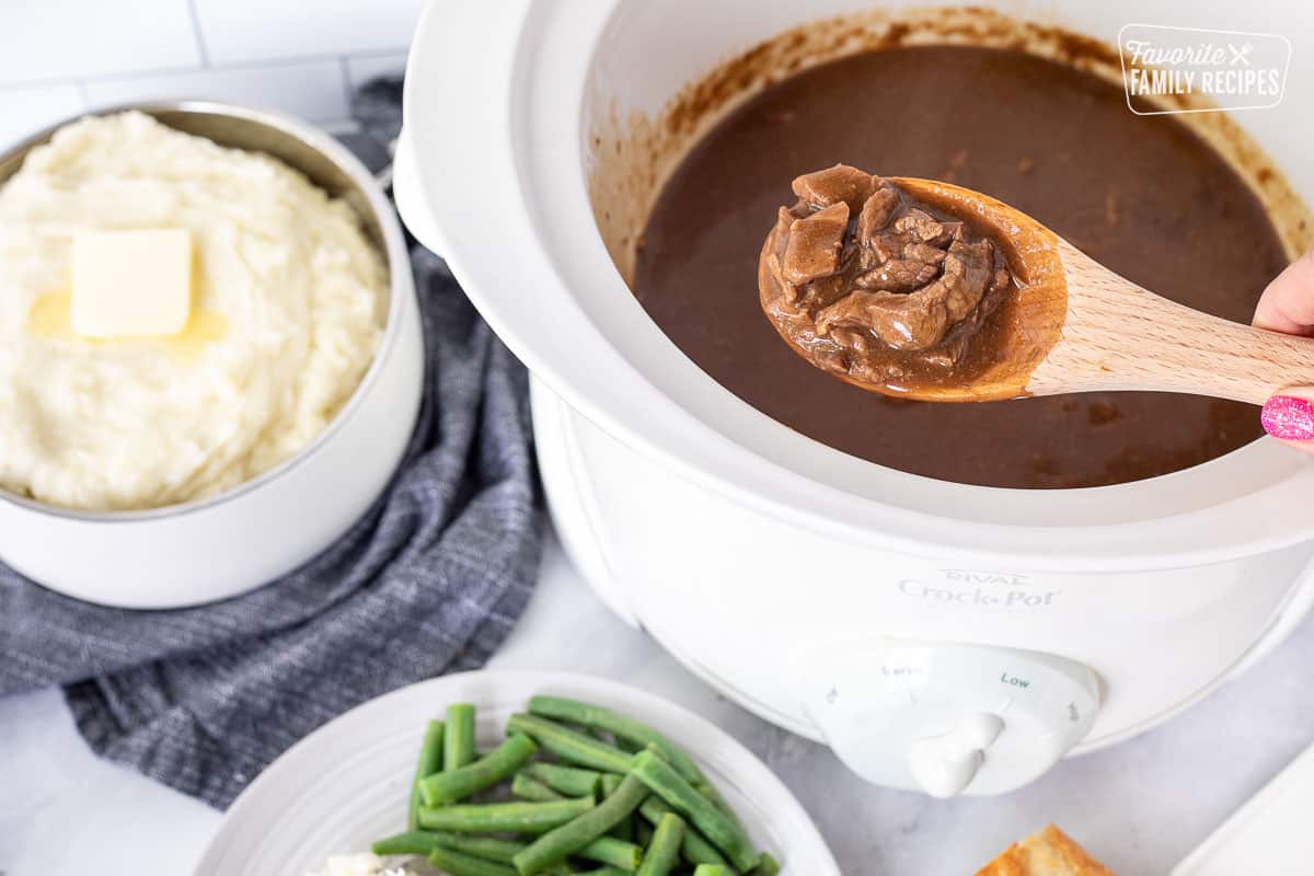 Wooden spoon with Crockpot Steak and Gravy next to a bowl of mashed potatoes and plate of green beans.
