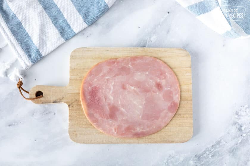 Cutting board with a slice of ham for Ham Roll Up with Pickle.