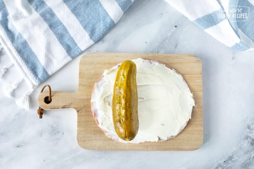 Cutting board with a slice of ham covered in cream cheese and a pickle on top.