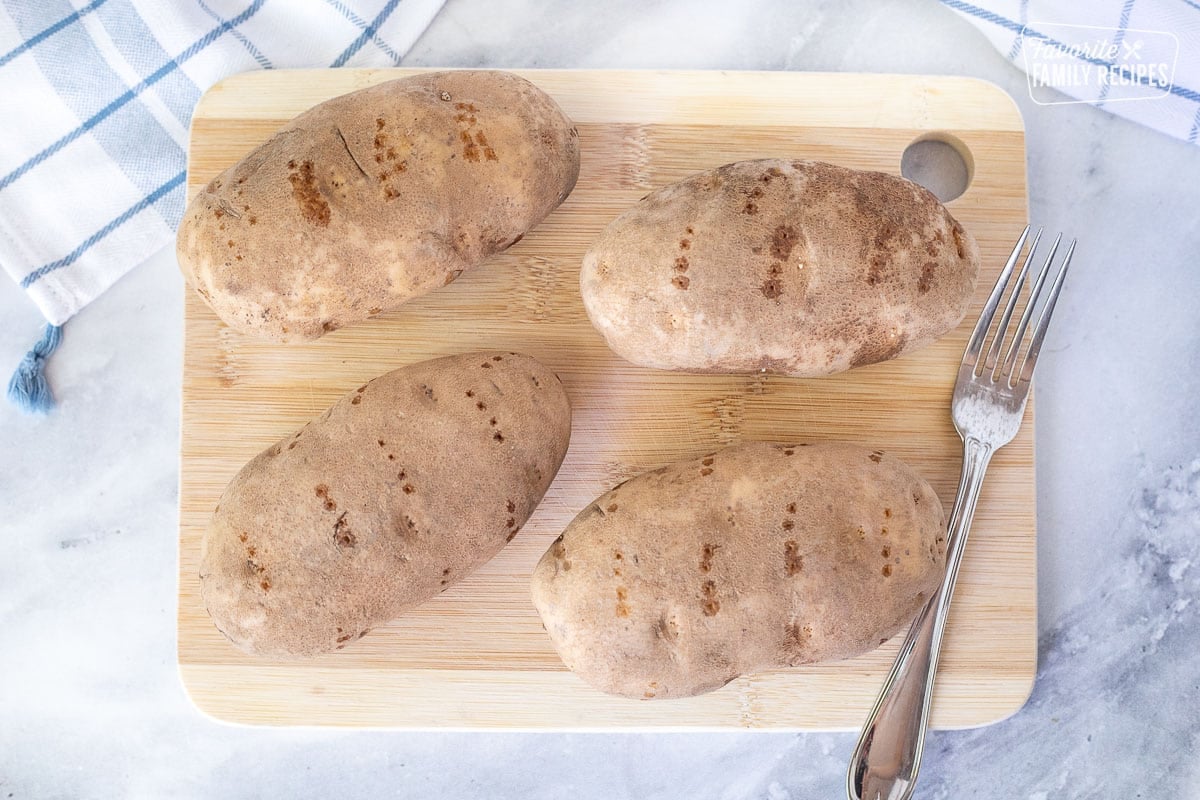 Cutting board with four washed potatoes poked with a fork. Fork on the side.