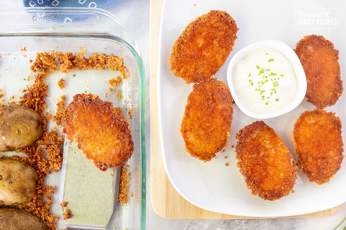 Spatula holding a Parmesan Crusted Potato in a baking dish next to a platter of Parmesan Crusted Potatoes with sour cream and chives.