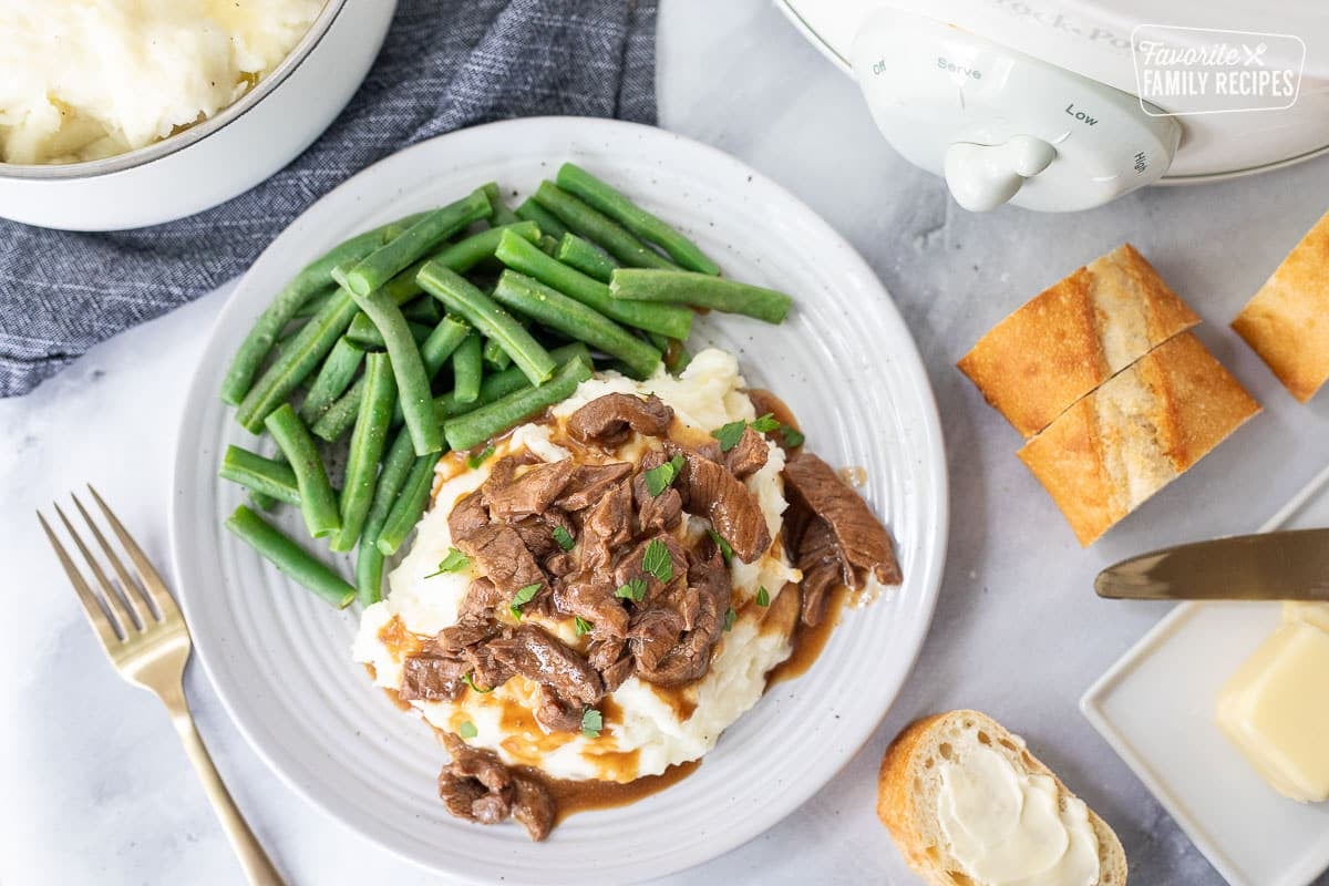 Plate with mashed potatoes covered with Crockpot Steak and Gravy and green beans on the side. Sliced baguette with butter on the side.