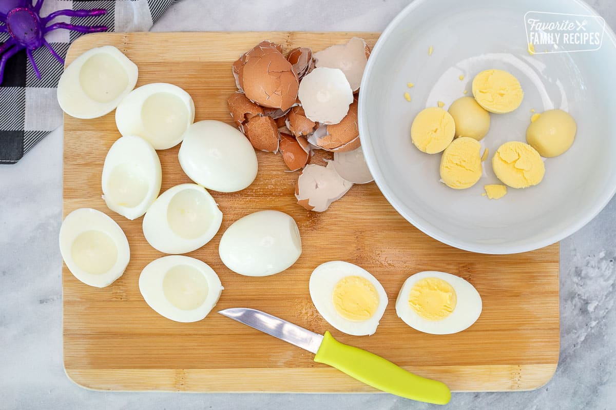 Cutting board with hard boiled eggs sliced half with the yolks taken out and placed in a bowl for Halloween Deviled Eggs.