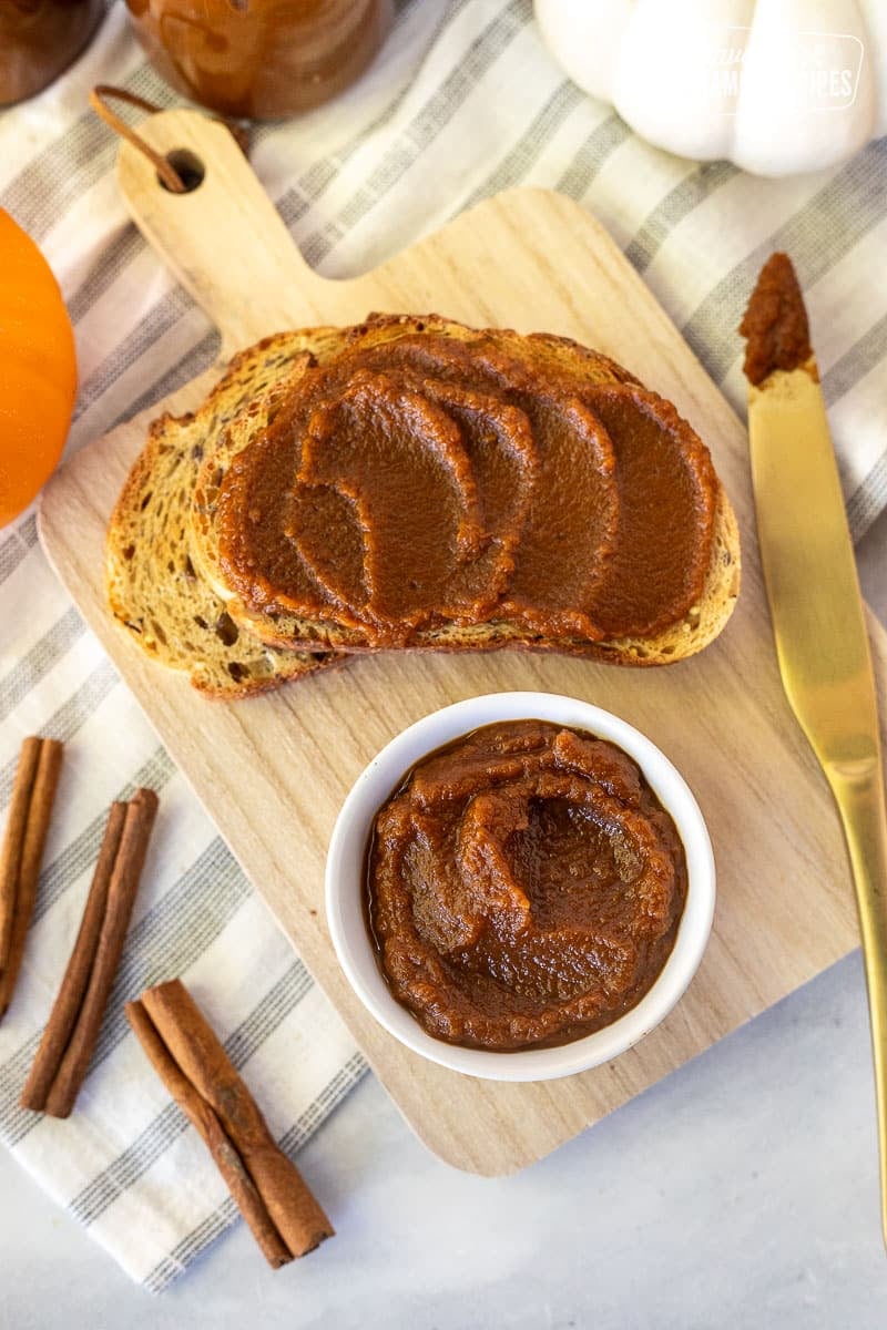 Maple Pumpkin Butter Spread is about to change your life. It is so yummy and easy to make! Perfect for spreading on dinner rolls hot out of the oven. Small bowl of Pumpkin Butter on a cutting board with toasted bread.