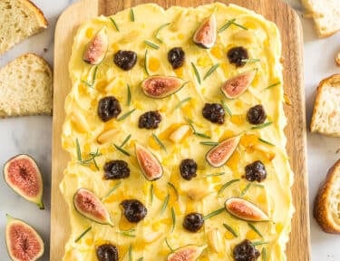 A butter board surrounded by bread slices, rosemary, and a bowl of honey.