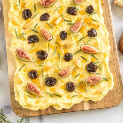 A butter board surrounded by bread slices, rosemary, and a bowl of honey.